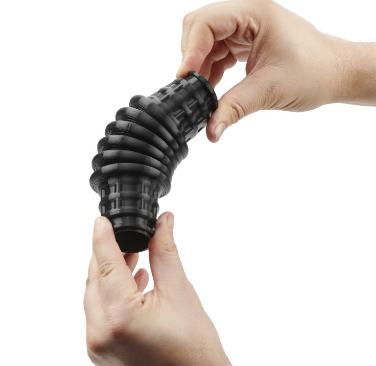 Pièce noire souple imprimée en 3D, déformée entre deux mains — Stratasys