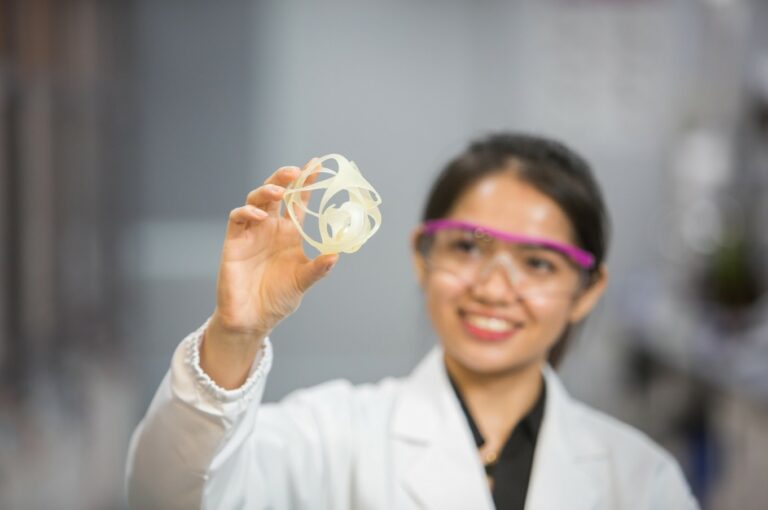 A researcher wearing a lab coat and safety glasses examines a white circular part