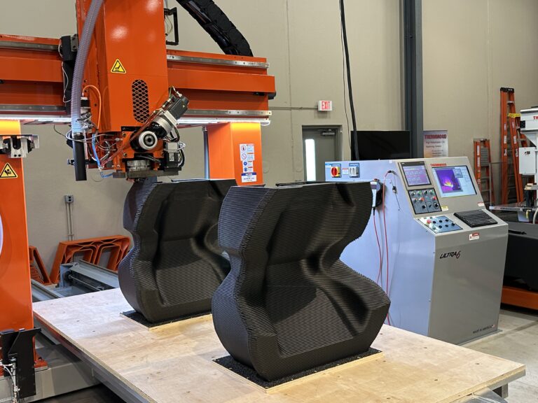 Thermwood extrusion head in front of two large printed parts and a control console