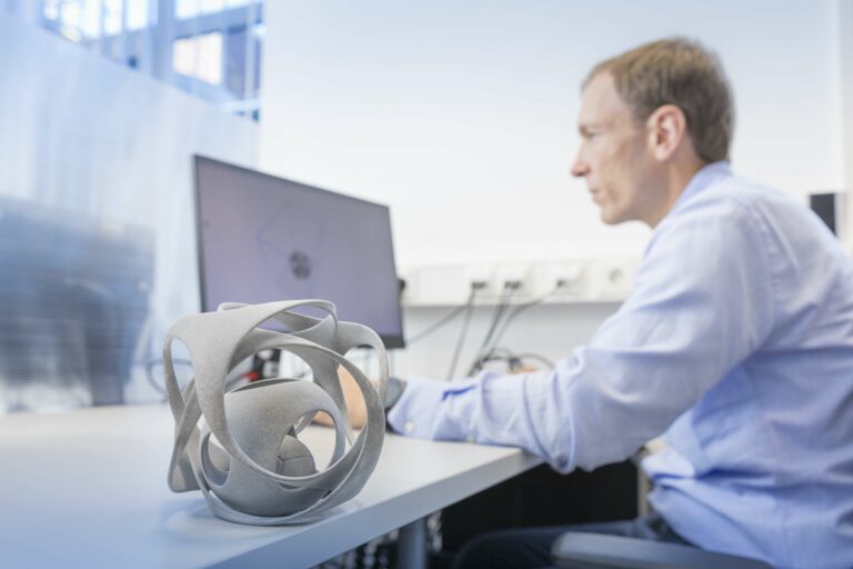 3D-printed part in the foreground with a person working at a computer in a laboratory