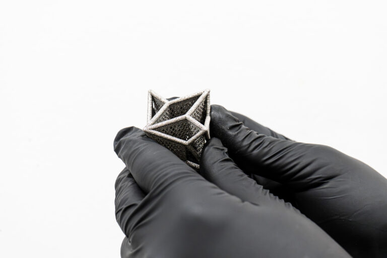 A gloved hand holds a small metal component with an open structure.