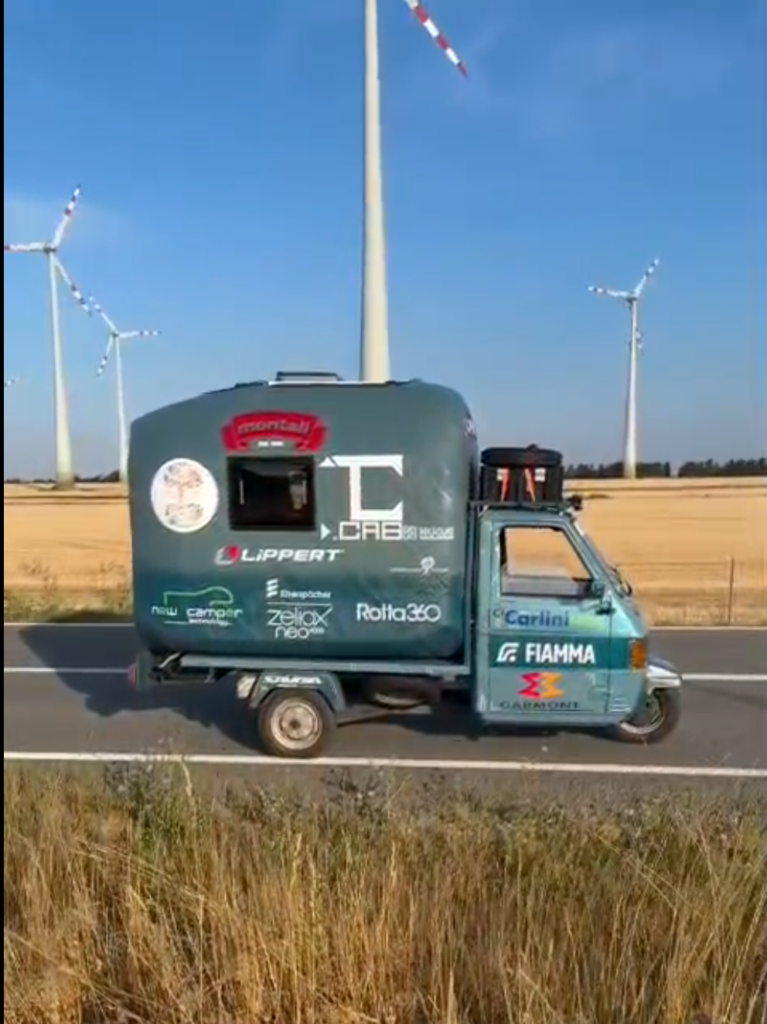 Piaggio Ape carrying the NUGAE camper module beside wind turbines.