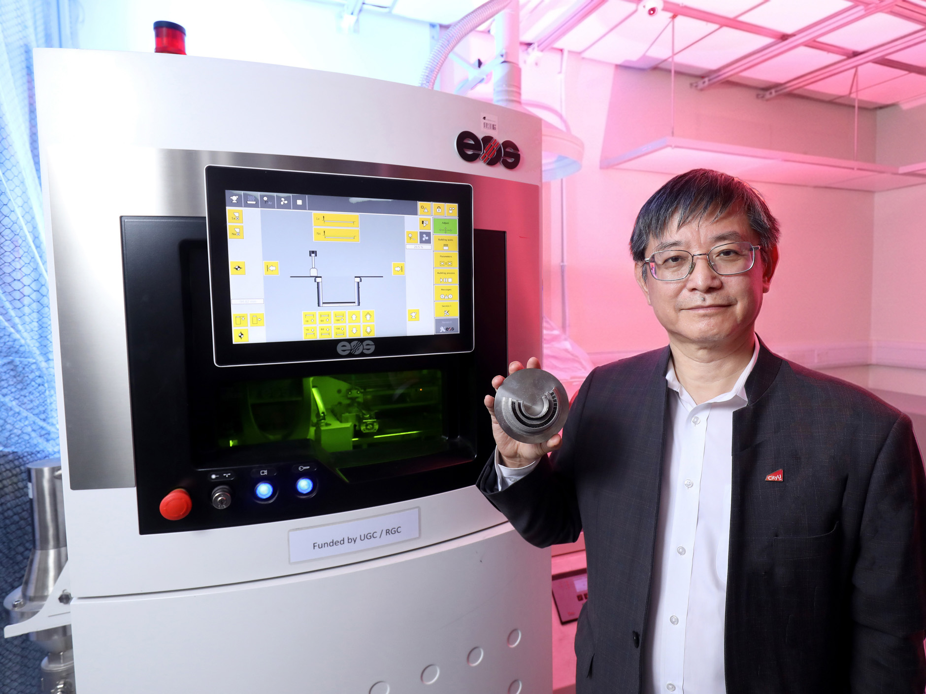 Professor Lu holding a metal component beside an EOS additive manufacturing machine.